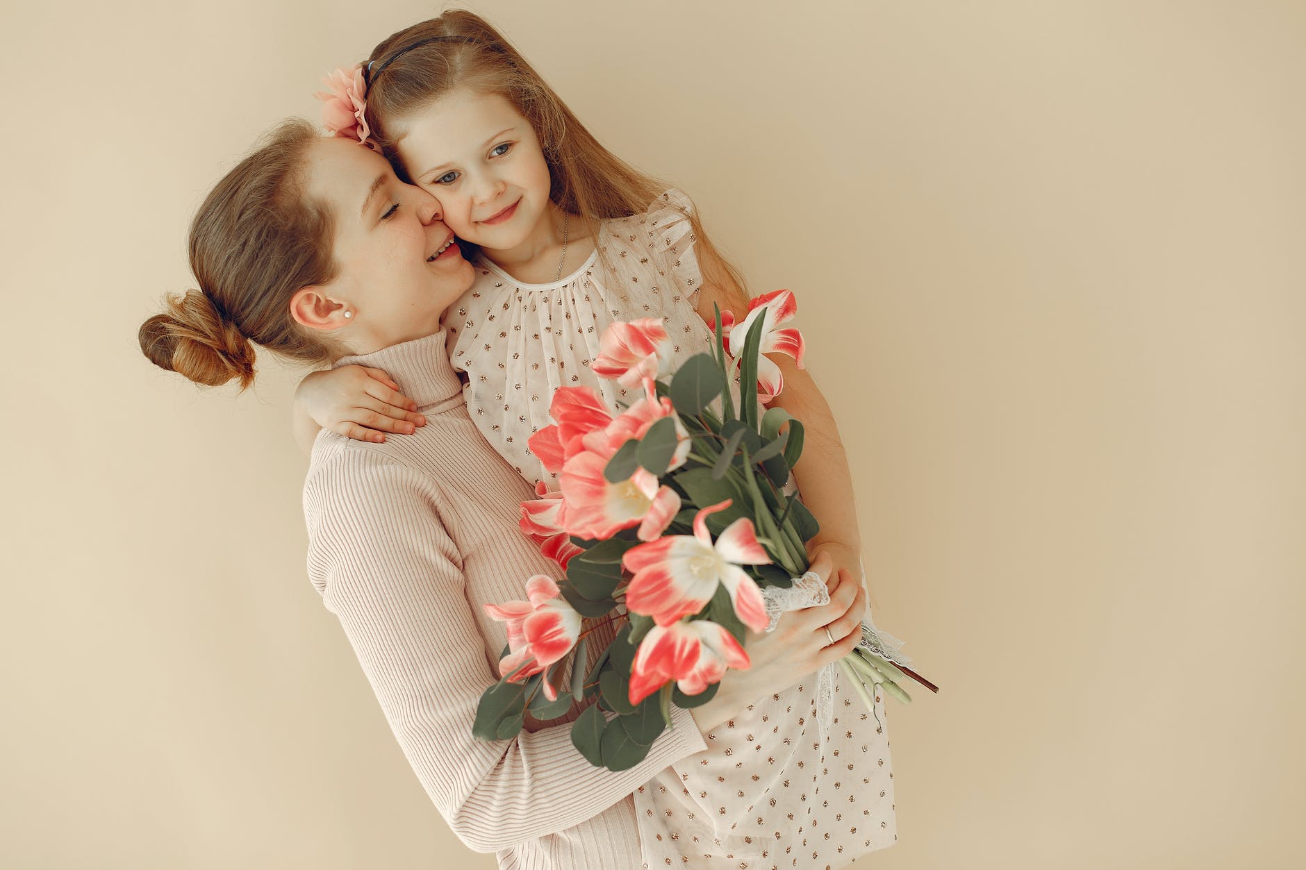 A mother presses her nose to her daughter's cheek, eyes closed. They stand in front of a pink background, holding matching pink flowers.