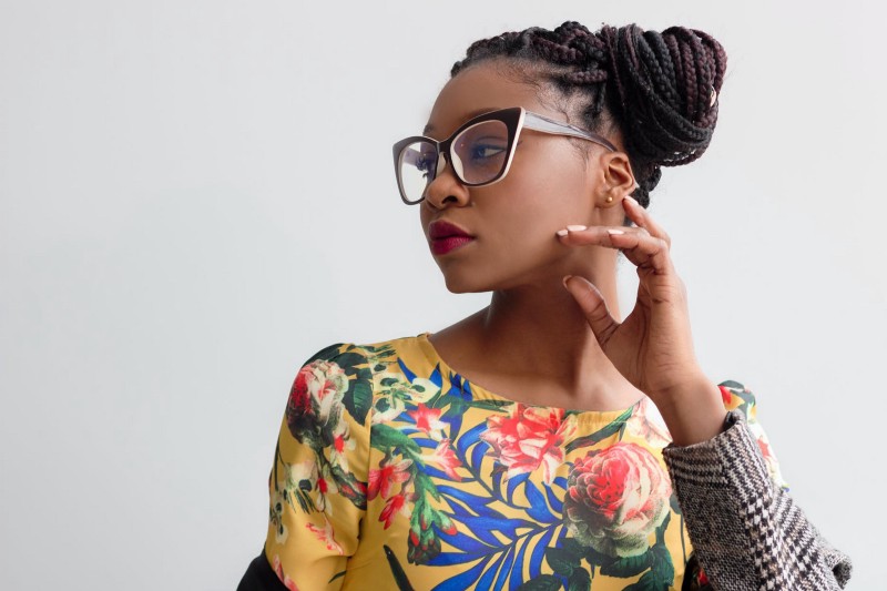 A young woman stares off to the side. Her hand grazes her beautiful face, and the bright colours of her shirt contrast with the plain, grey background. Her hair is pulled back in braids atop her head. She wears stylish glasses, and her lips are stained red. Classy.