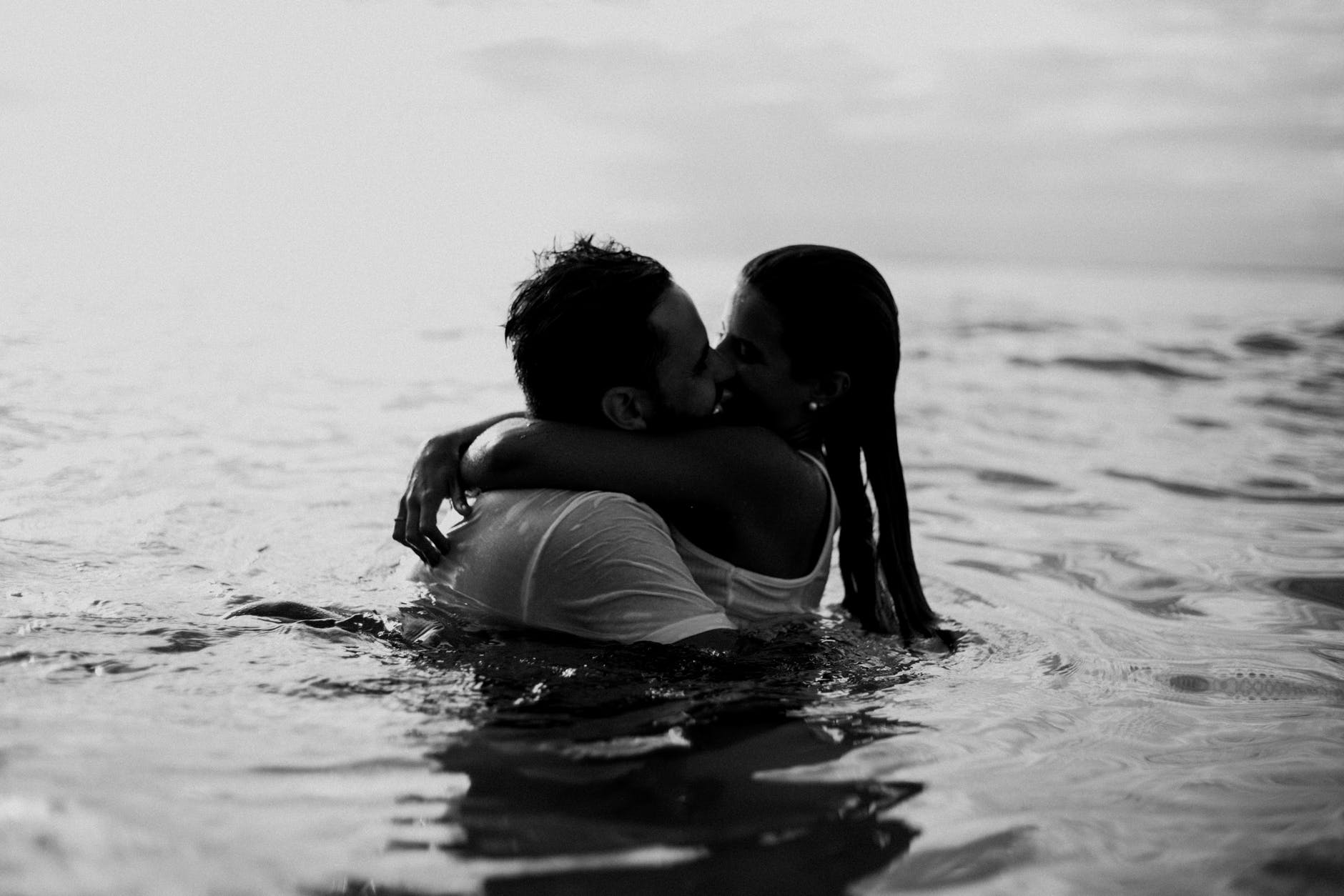 Black and white photo of a couple kissing in the water. They are both dressed in white, soaked, and look joyful.