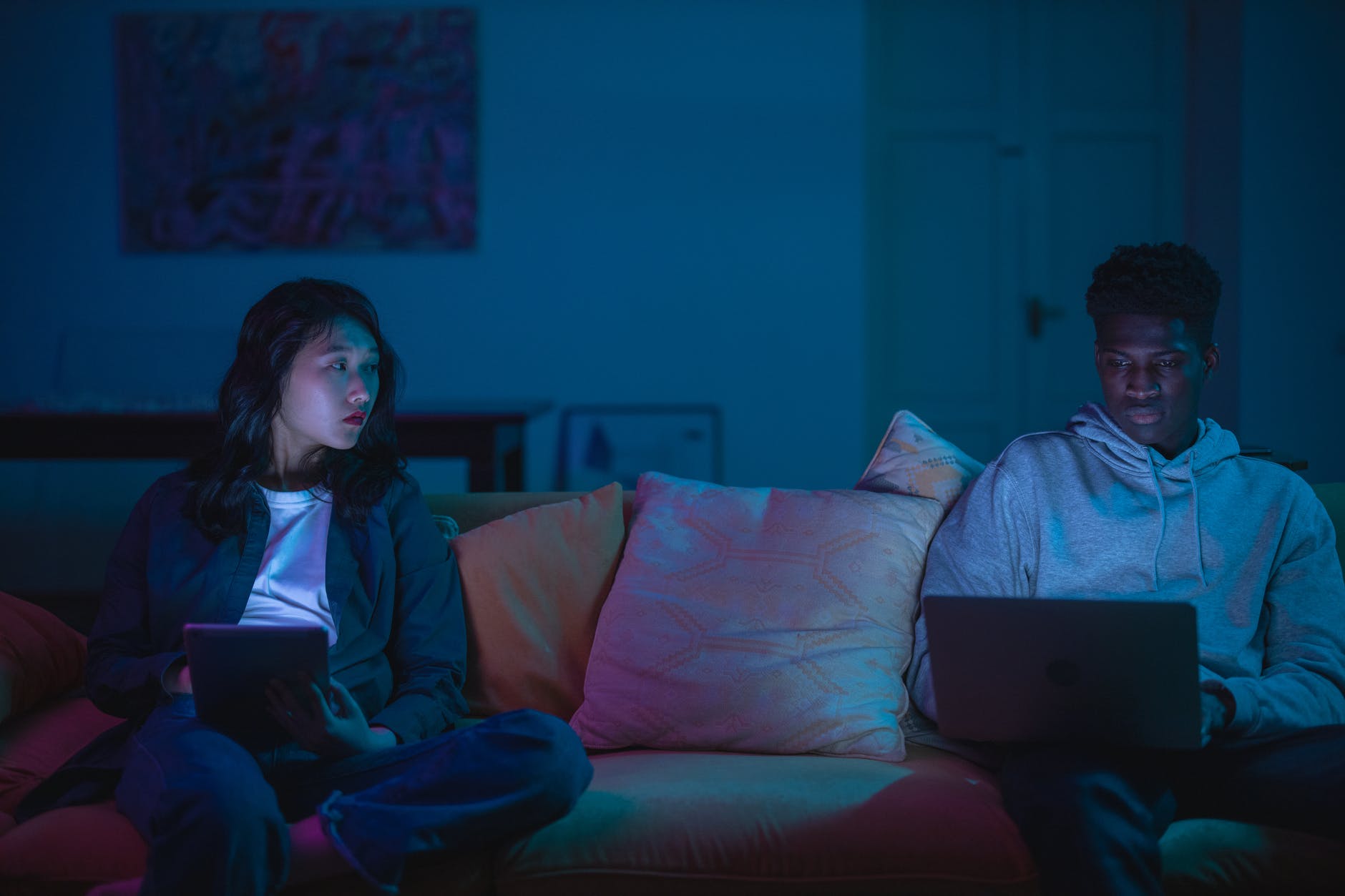 A couple is sitting on a couch, holding different devices. The long-haired womxn on the right is staring at her partner, with a concerned look on their face. Their partner is oblivious, looking at the screen of their computer.