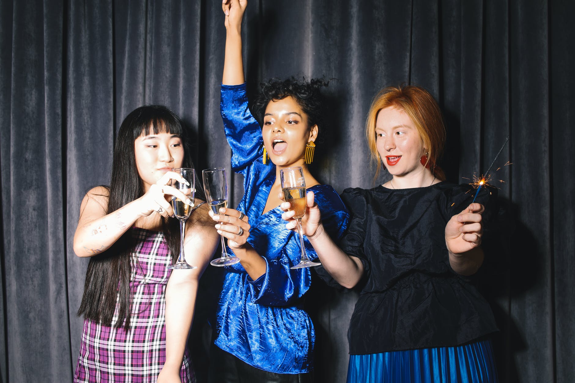 Three womxn stand side-by-side in front of a dark, velvet curtain, they cheers with champagne glasses and sparklers, wearing different celebratory expressions! 