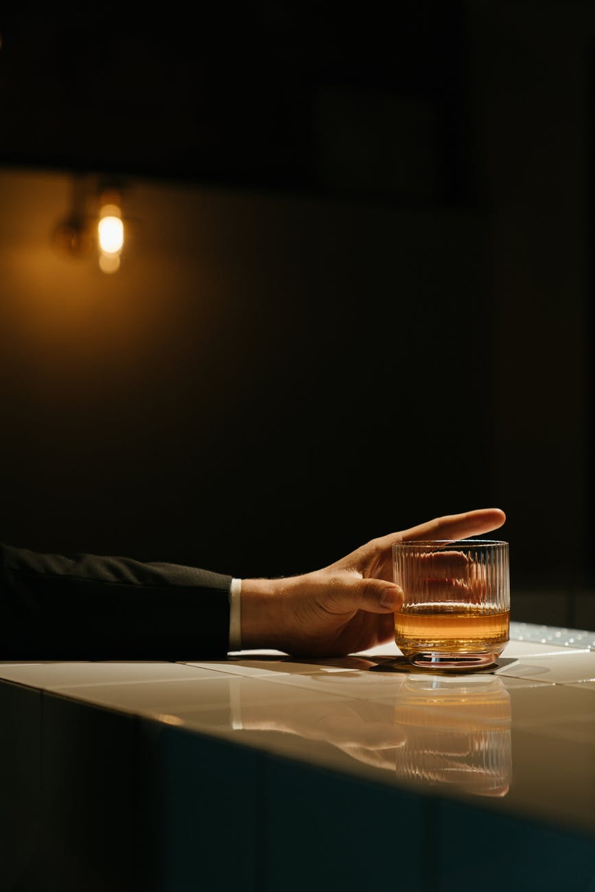 Reaching from off-view is a man's arm in a suit jacket, holding onto a glass of whiskey, neat.