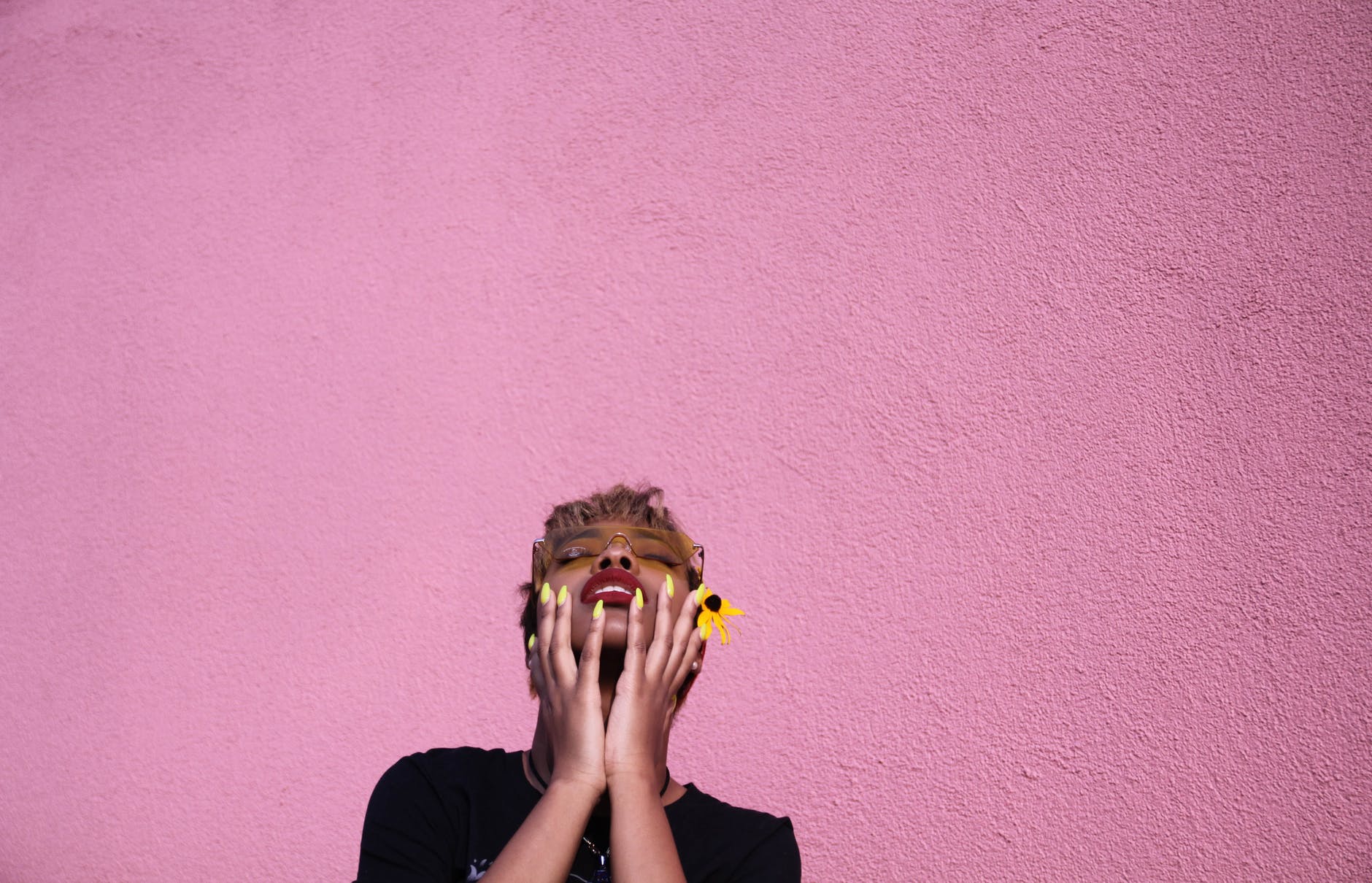 Pink background, a woman with short hair embraces her face. She has long, yellow nails and a sunflower.