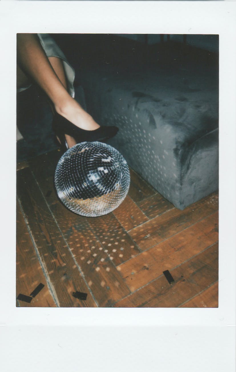 Polaroid picture of a suede couch and a woman's leg. She wears a silver dress and black high heels, and is resting one on a disco ball. 