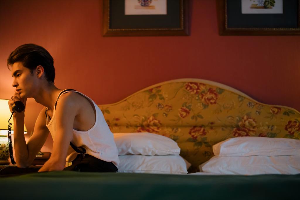 A man sits on the edge of a bed - green blanket, white pillows and yellow headboard. He has a white tank top on, dark pants and suspenders. He holds a black phone to his ear; expression stoic.